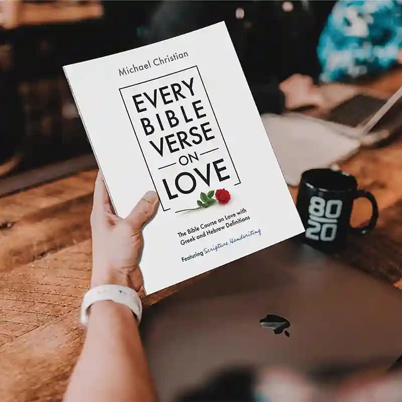 Image of a man's hand holding the book Every Bible Verse on Love over a laptop computer with a coffee cup on the side